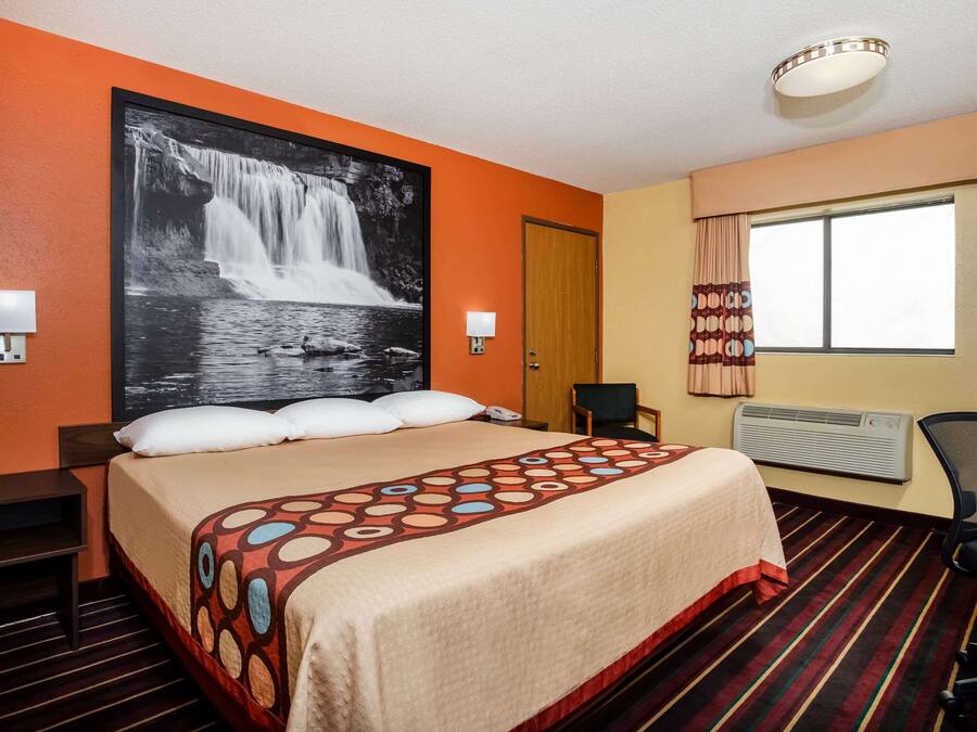 A hotel room with a large bed, a wall-mounted TV, a window with curtains, and a desk with a chair.