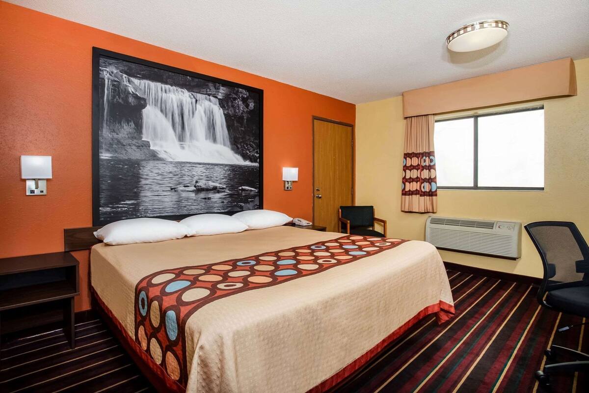 Ein Hotelzimmer mit Bett, einem großen Wandbild mit einem Wasserfall, einem Fenster mit Vorhängen, einem Schreibtisch und einem Stuhl.