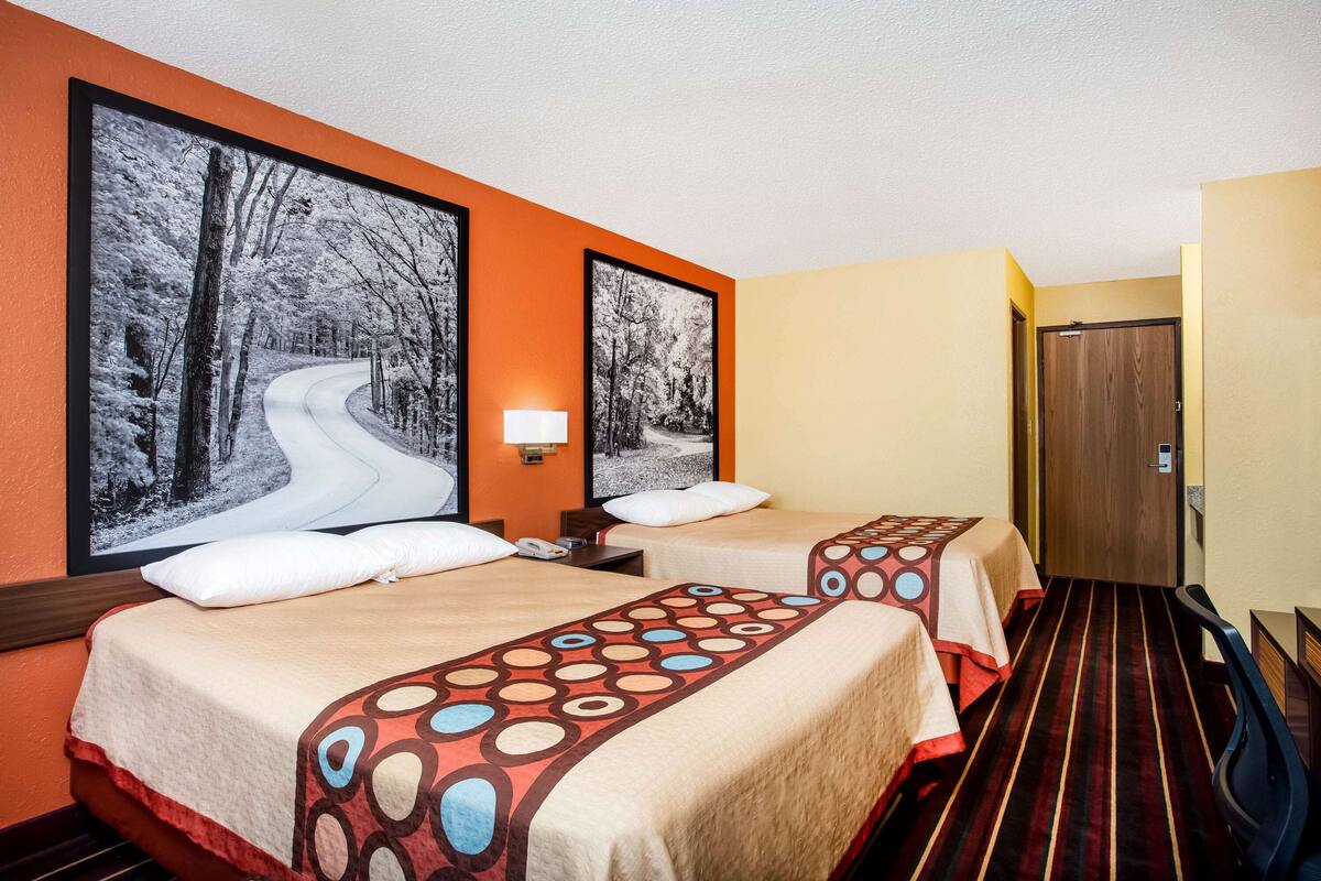 Ein Hotelzimmer mit zwei Betten, jeweils mit einem gemusterten Überwurf, einem Nachttisch mit Lampe und gerahmten Bildern an der Wand.
