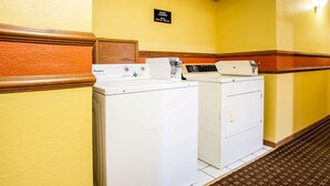 Une buanderie équipée de lave-linge et de sèche-linge Whirlpool, un panneau indiquant les heures d’ouverture de la buanderie pour les clients, et un banc en bois.