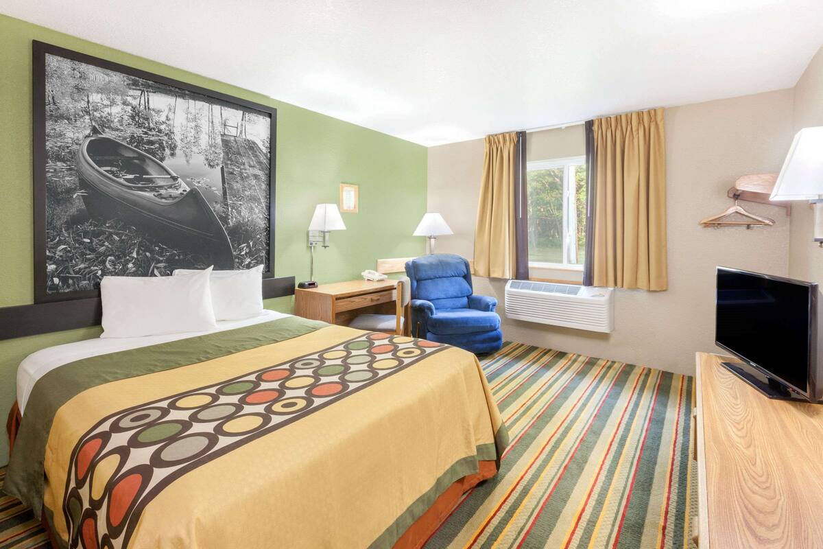Quarto de hotel com cama, escrivaninha, cadeira, televisão e janela com cortinas.
