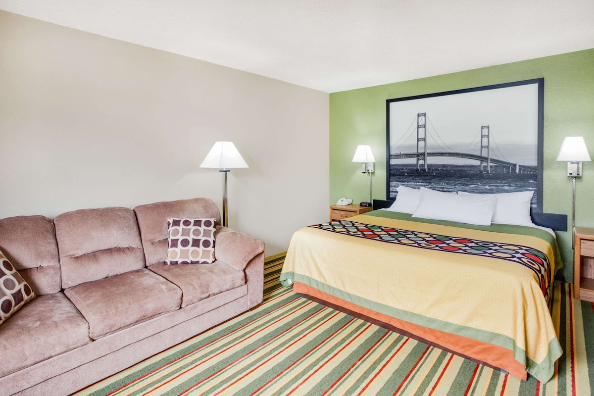 Quarto de hotel com cama, sofá, duas luminárias fixadas na parede, um quadro de uma ponte e um tapete listrado.