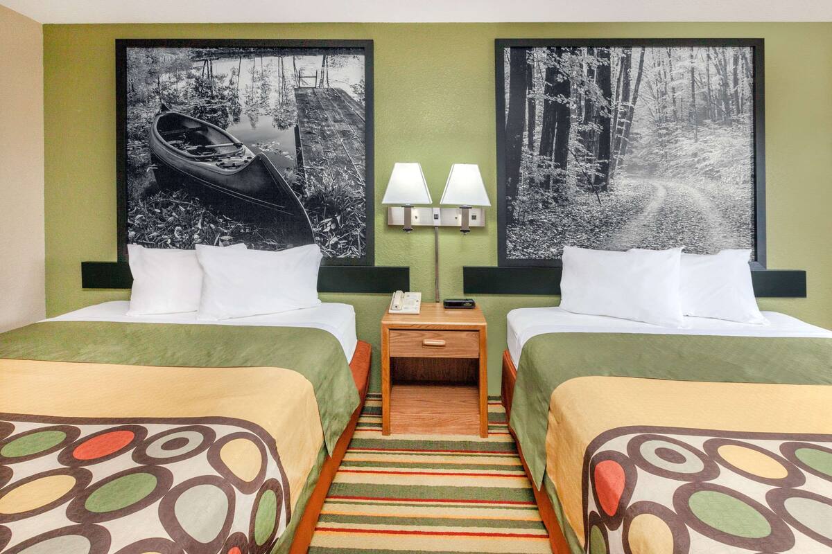 Quarto de hotel com duas camas, uma mesa de cabeceira com abajur e duas grandes molduras na parede.