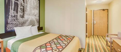 A hotel room with a bed, a desk, and a chair. There is a framed picture on the wall and a door to the right.