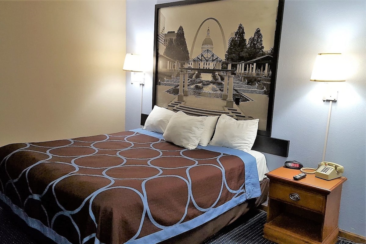 Une chambre d’hôtel avec un lit, une table de chevet avec une lampe, un téléphone et un tableau encadré au mur.
