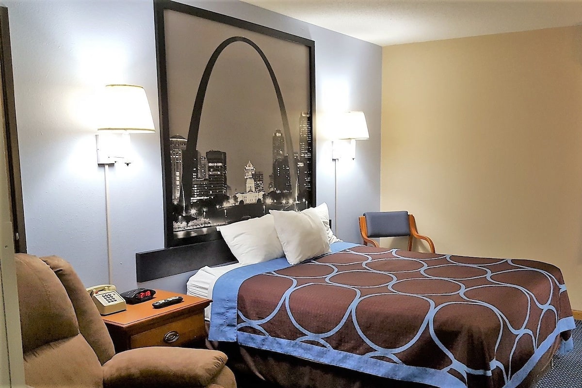 Une chambre d’hôtel avec un lit, deux lampes de chevet, une chaise, un bureau avec un téléphone et une grande affiche représentant un paysage urbain accrochée au mur.