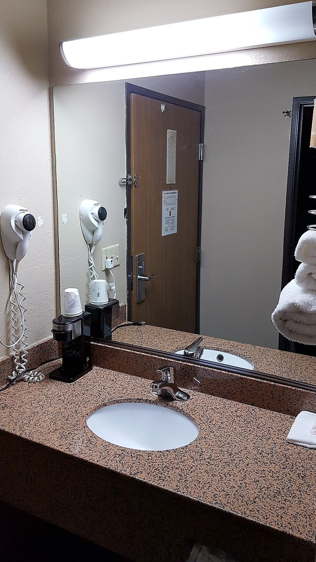 A bathroom with a granite countertop, a sink, a mirror, a hairdryer, a coffee maker, and a towel rack.