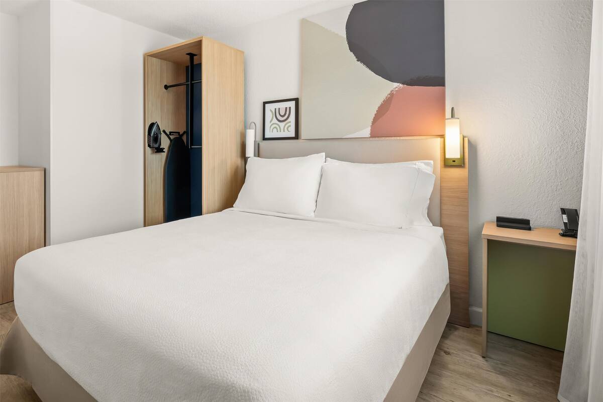 A hotel room with a large bed, a wooden wardrobe, a desk with a lamp, and a wall-mounted artwork.