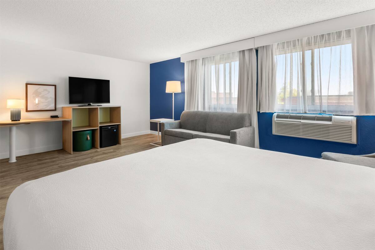 A hotel room with a large bed, a flat-screen TV, a desk with a lamp, a chair, and a window with curtains.
