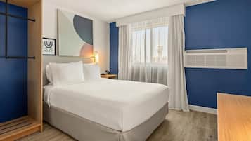 A hotel room with a bed, a bedside table, a lamp, a window with curtains, and an air conditioning unit.