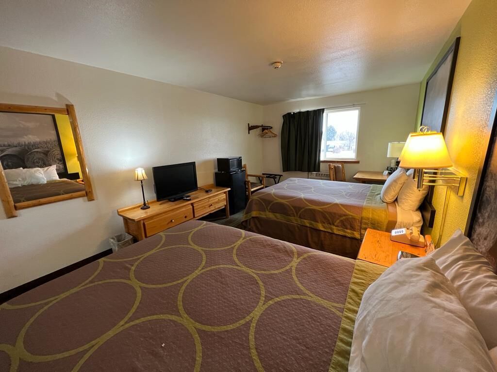 Ein Hotelzimmer mit zwei Betten, einem Schreibtisch mit Lampe, einem Fernseher und einem fensterbepflanzten Fenster.