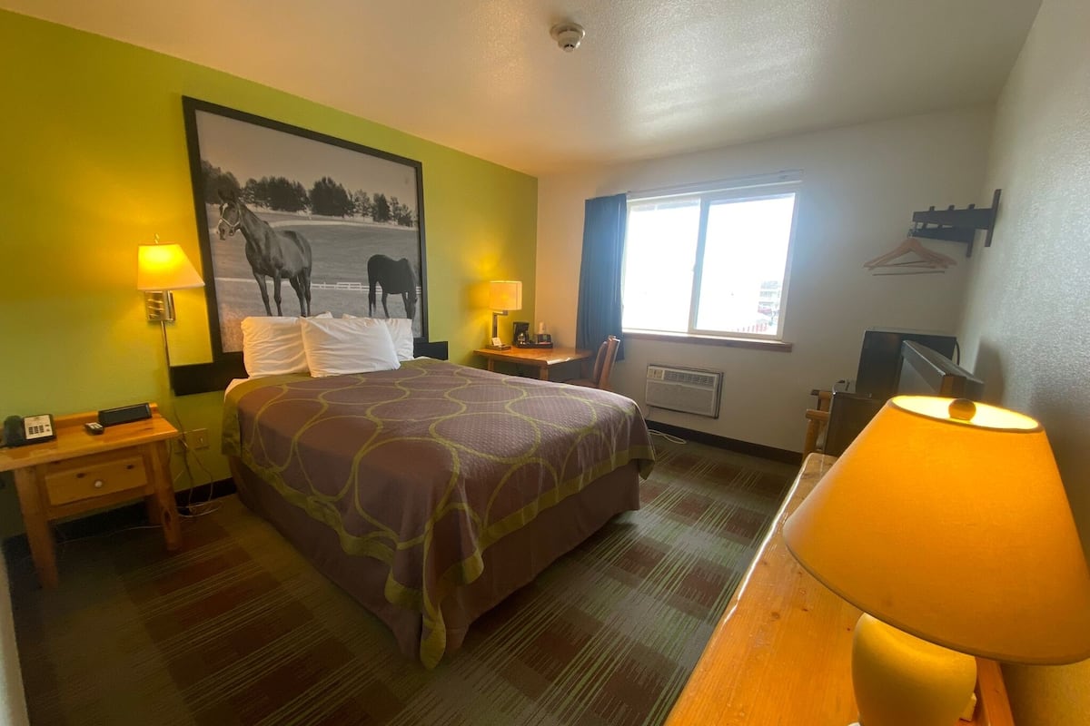 A hotel room with a bed, two bedside tables, a desk with a lamp, a window, and a wall-mounted TV.