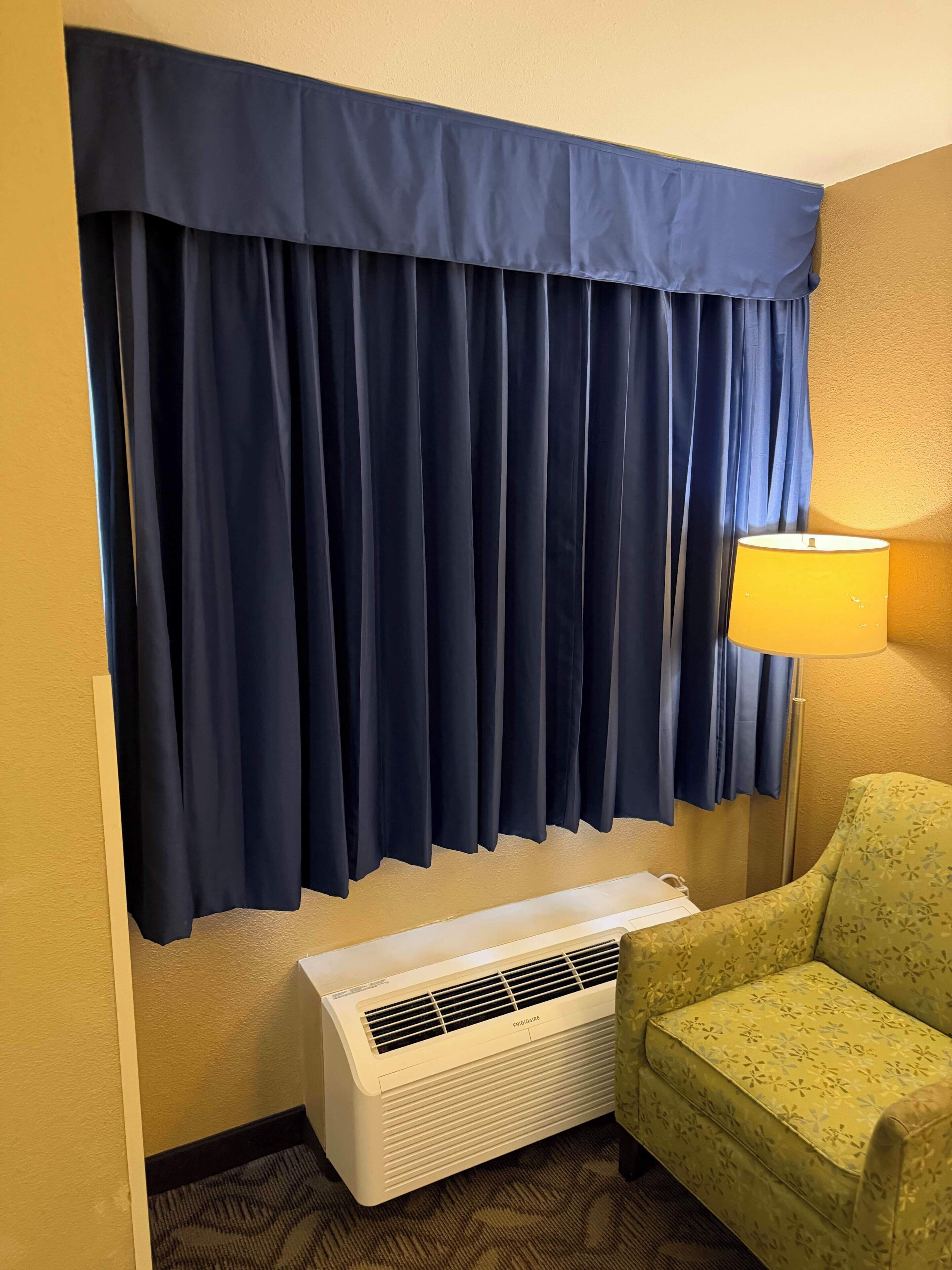 A room with a large window covered by dark blue curtains, a standing heater, and a green armchair.