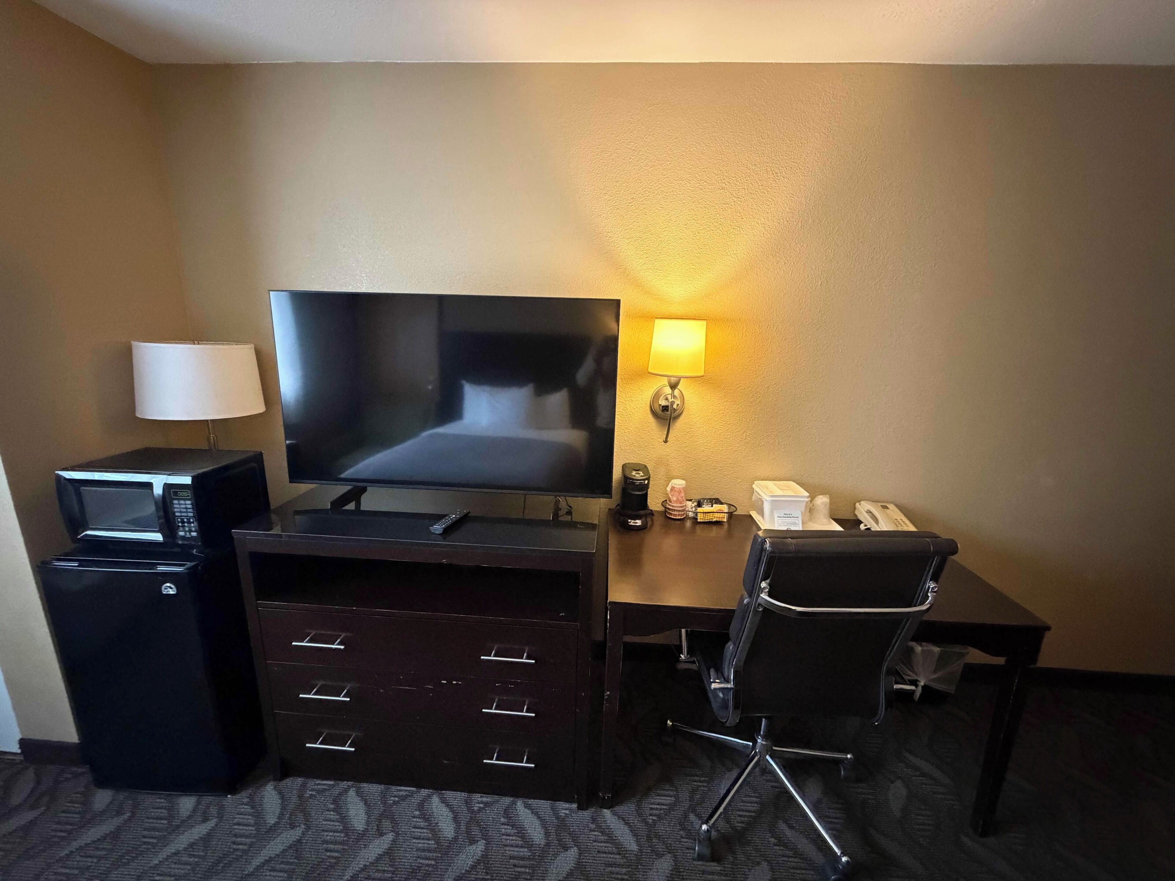 Een hotelkamer met een bed, een televisie, een bureau met een lamp, een magnetron, een koelkast en een stoel.
