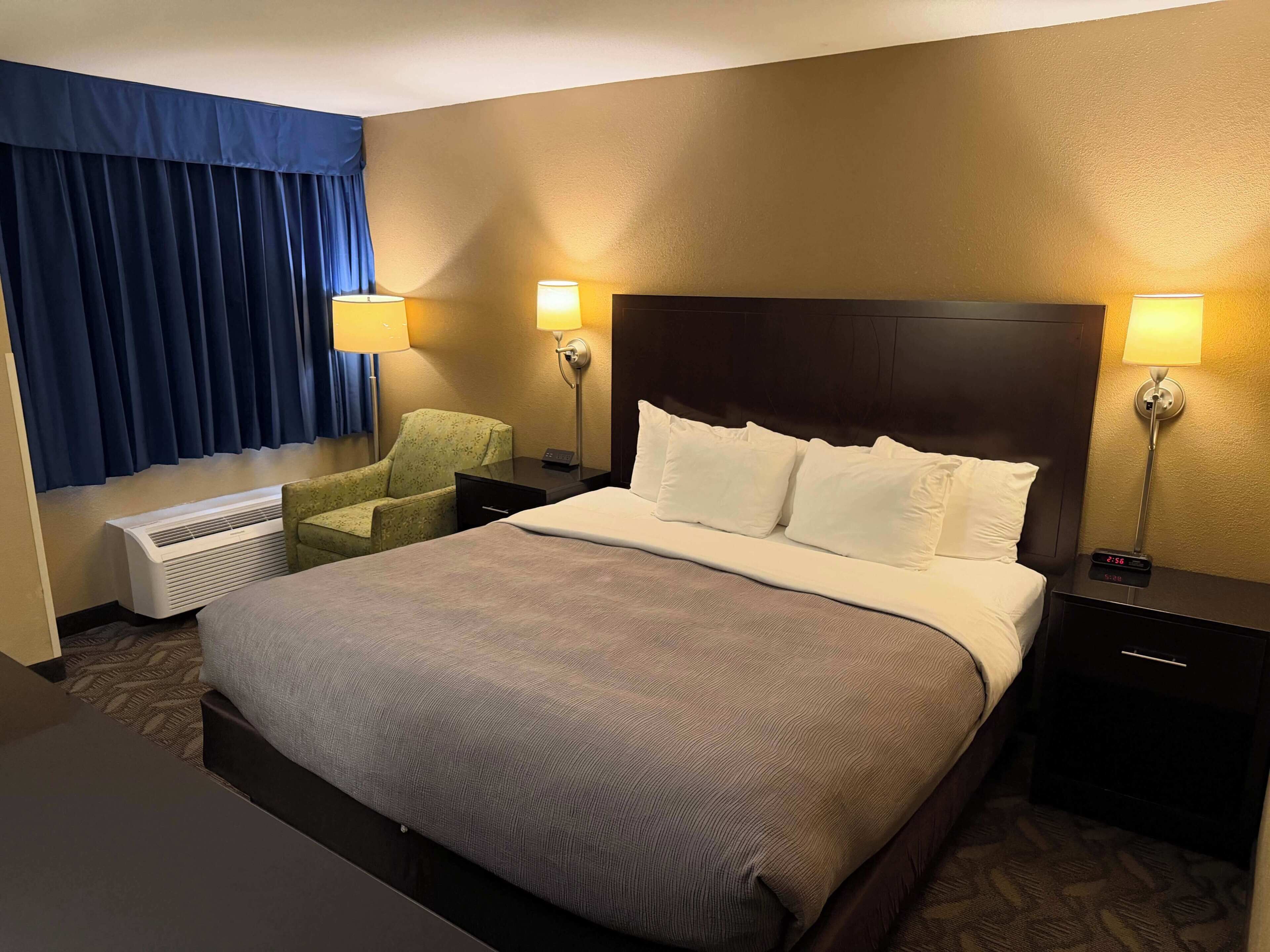 A hotel room with a large bed, two bedside lamps, a chair, a heater, and a window with curtains.