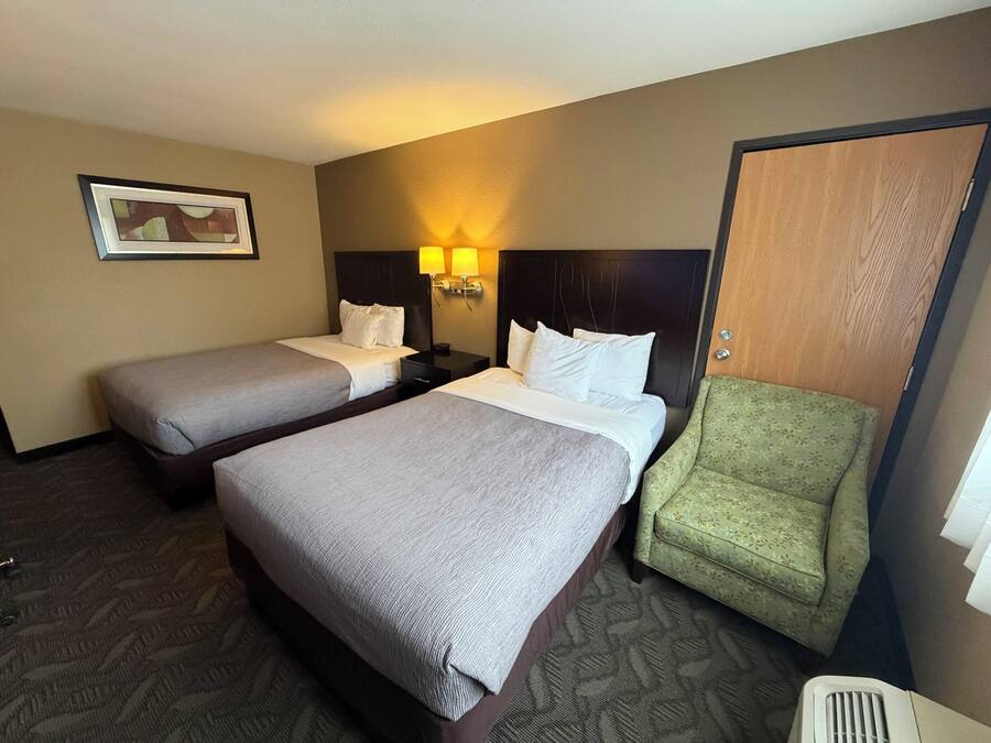 A hotel room with two beds, a green armchair, a nightstand with a lamp, and a framed picture on the wall.
