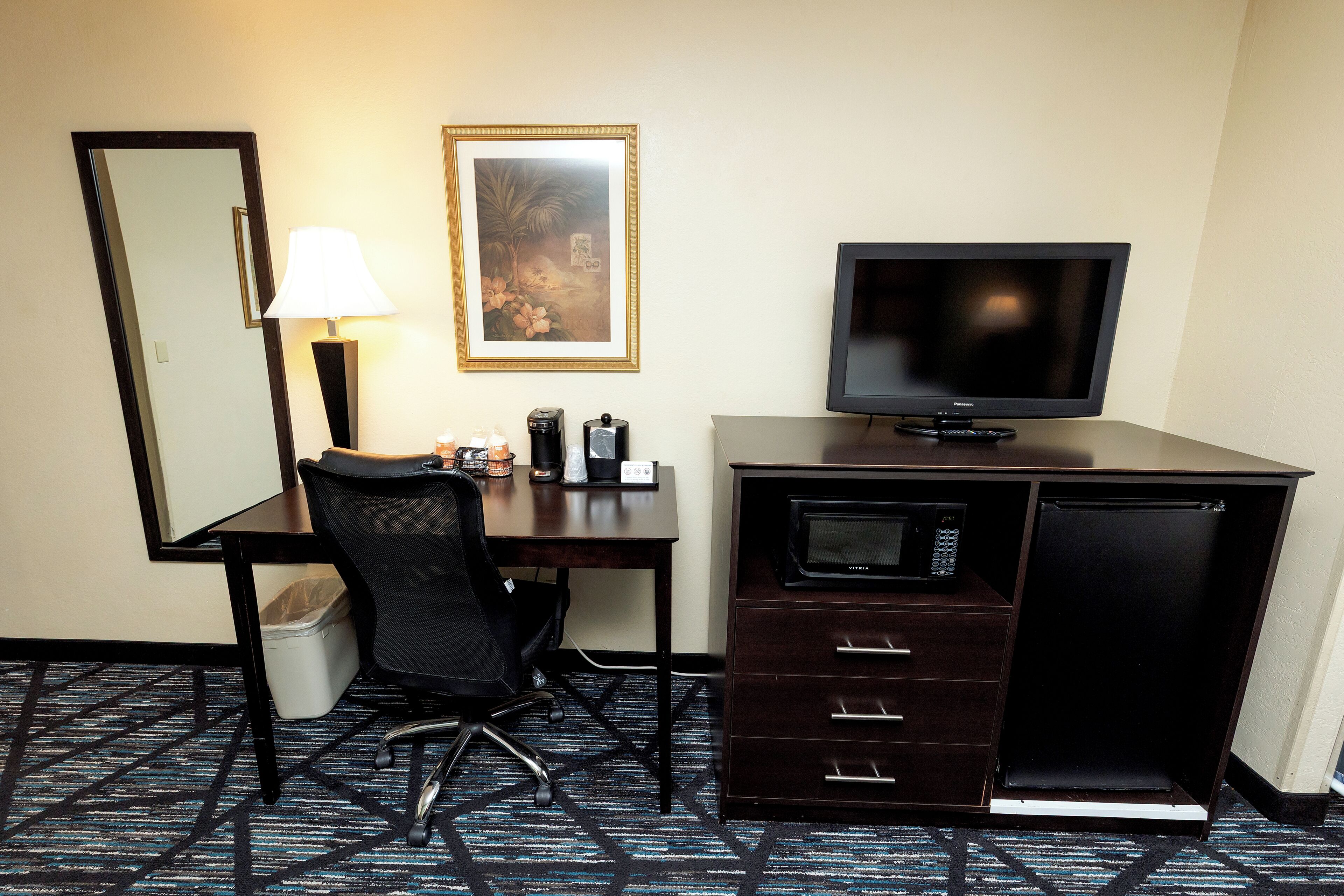 A hotel room with a desk, chair, television, microwave, and a trash bin.