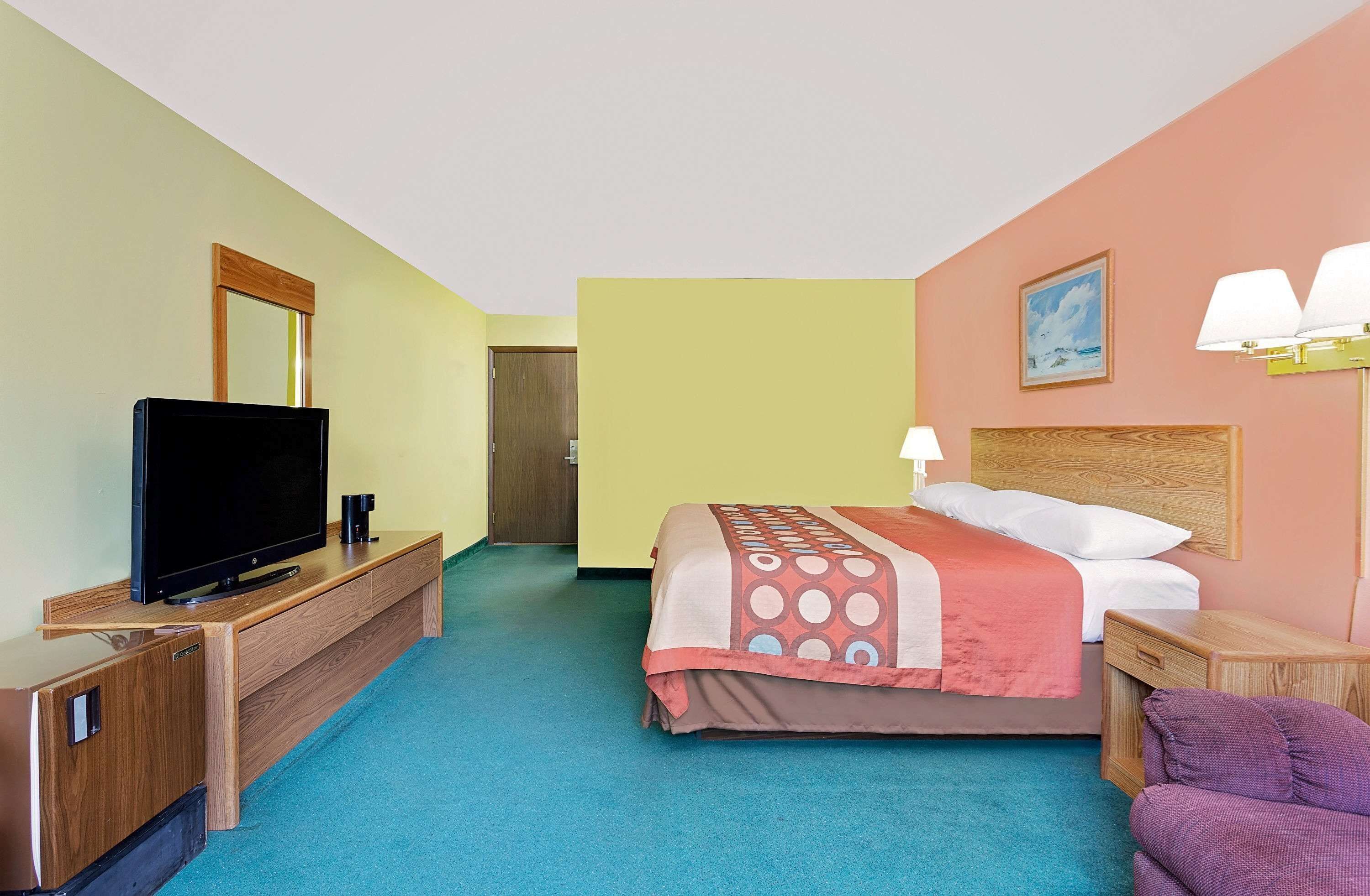 Habitación de hotel con una cama, un televisor sobre un mueble de madera, una mesita de noche, una lámpara y un cuadro en la pared.