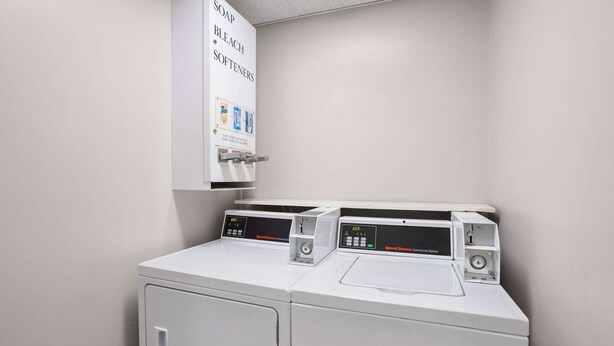 A laundry area with two front-loading washing machines and a wall-mounted dispenser for soap, bleach, and softeners. - Super 8 by Wyndham Henrietta/Rochester Area (Henrietta)