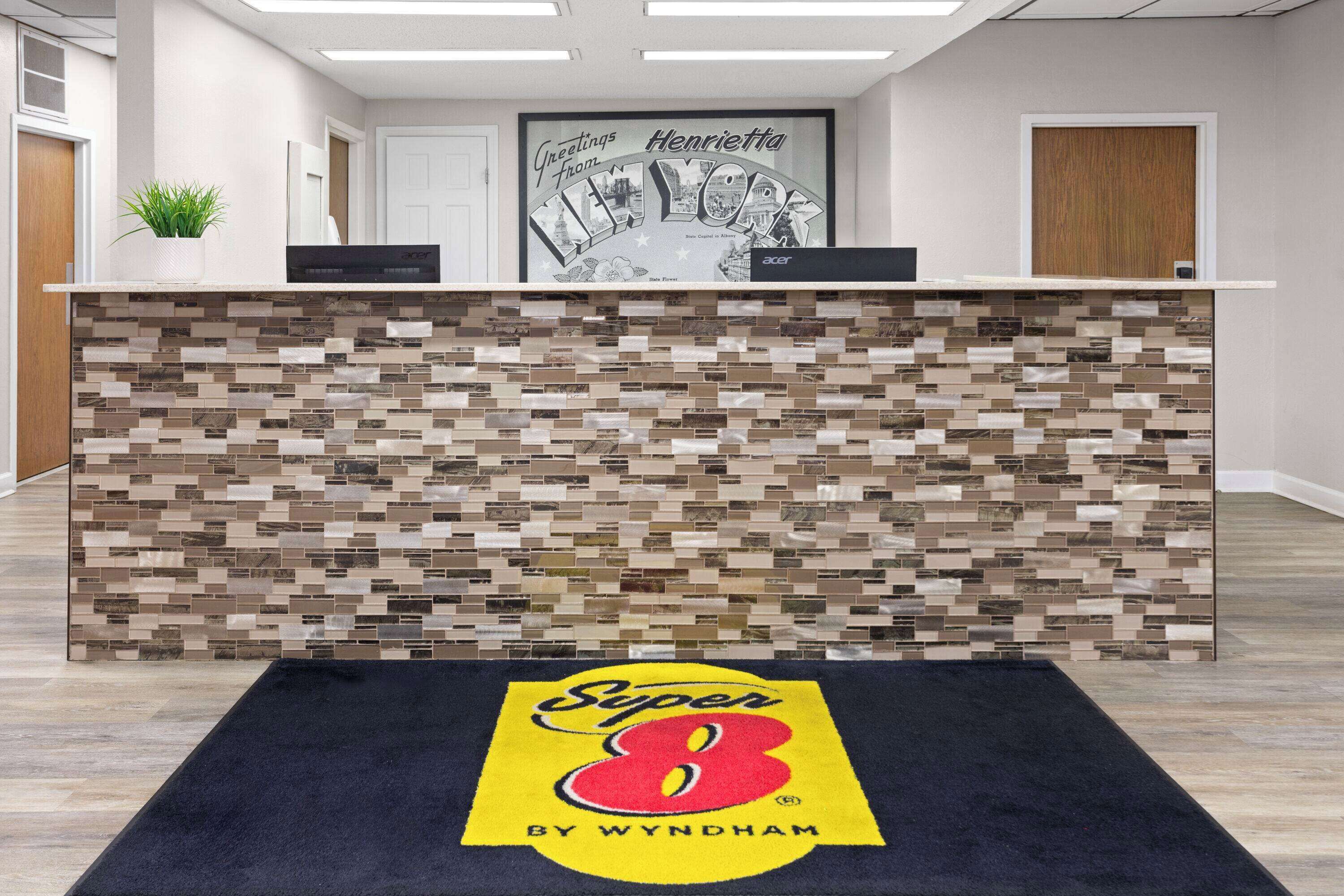 A reception area with a mosaic tile wall, a framed 'Greetings from New York' sign, and a 'Super 8 by Wyndham' rug.
