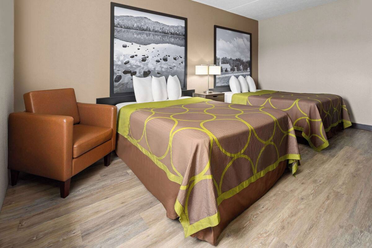 Habitación de hotel con dos camas, una colcha de tonos marrón y amarillo, un sillón de cuero marrón y dos grandes cuadros de paisajes en la pared.