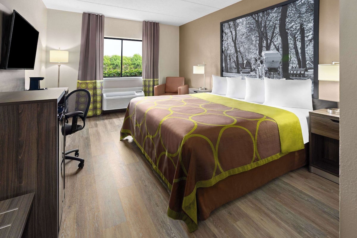 Habitación de hotel con cama, escritorio, silla, televisor y ventanas con cortinas.