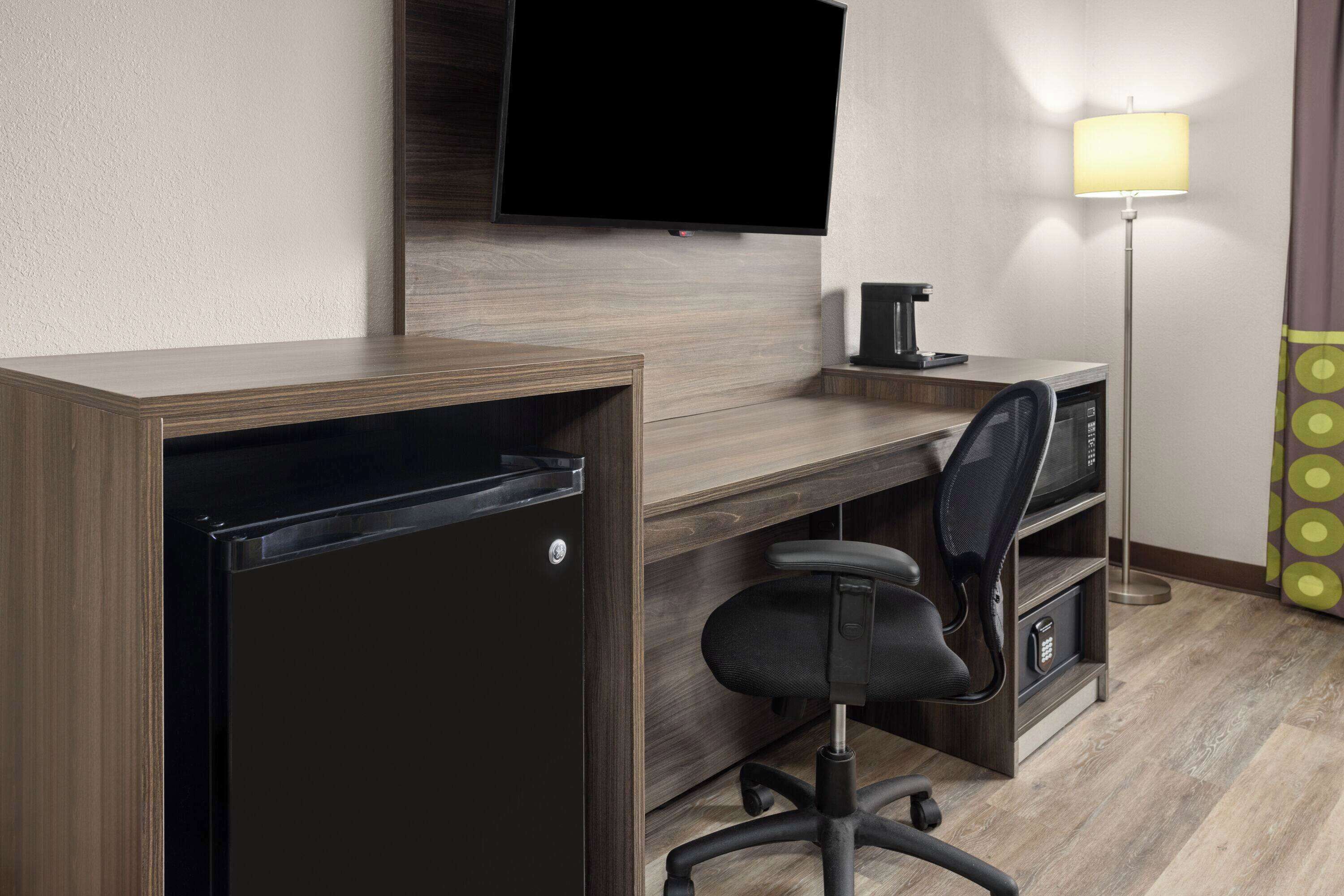 A hotel room with a wooden desk, a black office chair, a flat-screen TV mounted on the wall, a floor lamp, and a minibar.