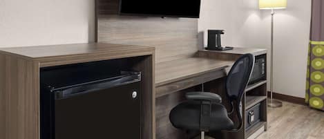 A hotel room with a wooden desk, a black office chair, a flat-screen TV mounted on the wall, a floor lamp, and a minibar.
