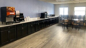 Cafetería con una barra larga, máquinas de café y un dispensador de fruta. Hay mesas y sillas para comer.