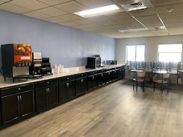 Uma cafeteria com um balcão comprido, máquinas de café e um dispensador de frutas. Há mesas e cadeiras para refeições.