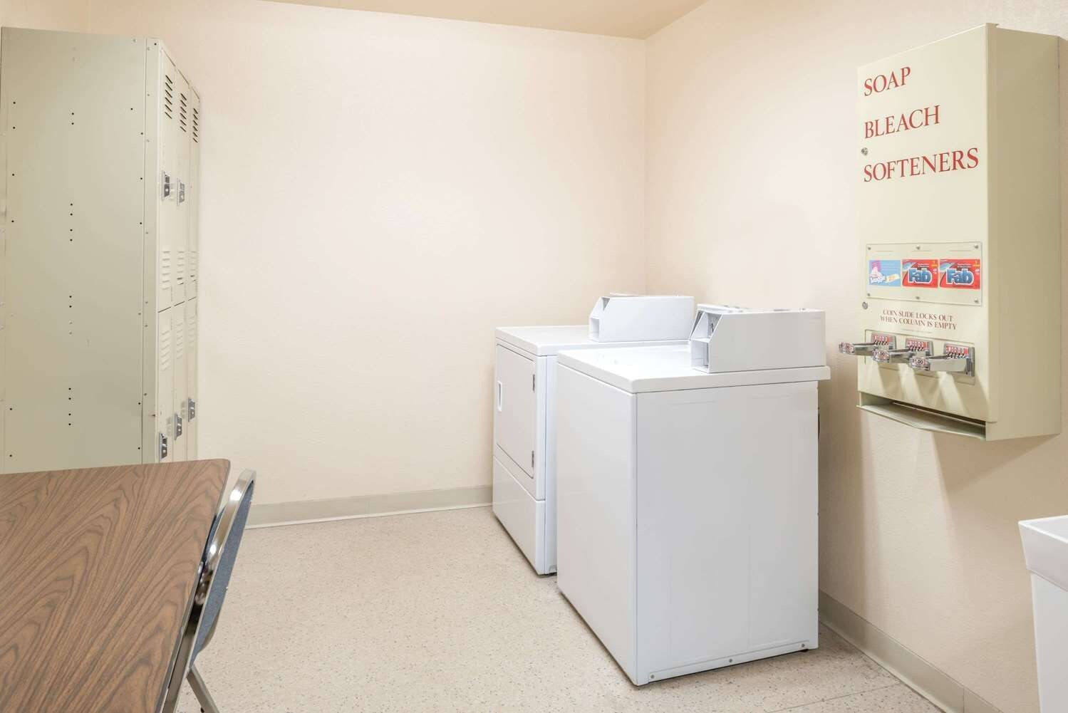 A laundry room with two white washers and dryers, a coin-operated soap and bleach dispenser, and a metal locker.
