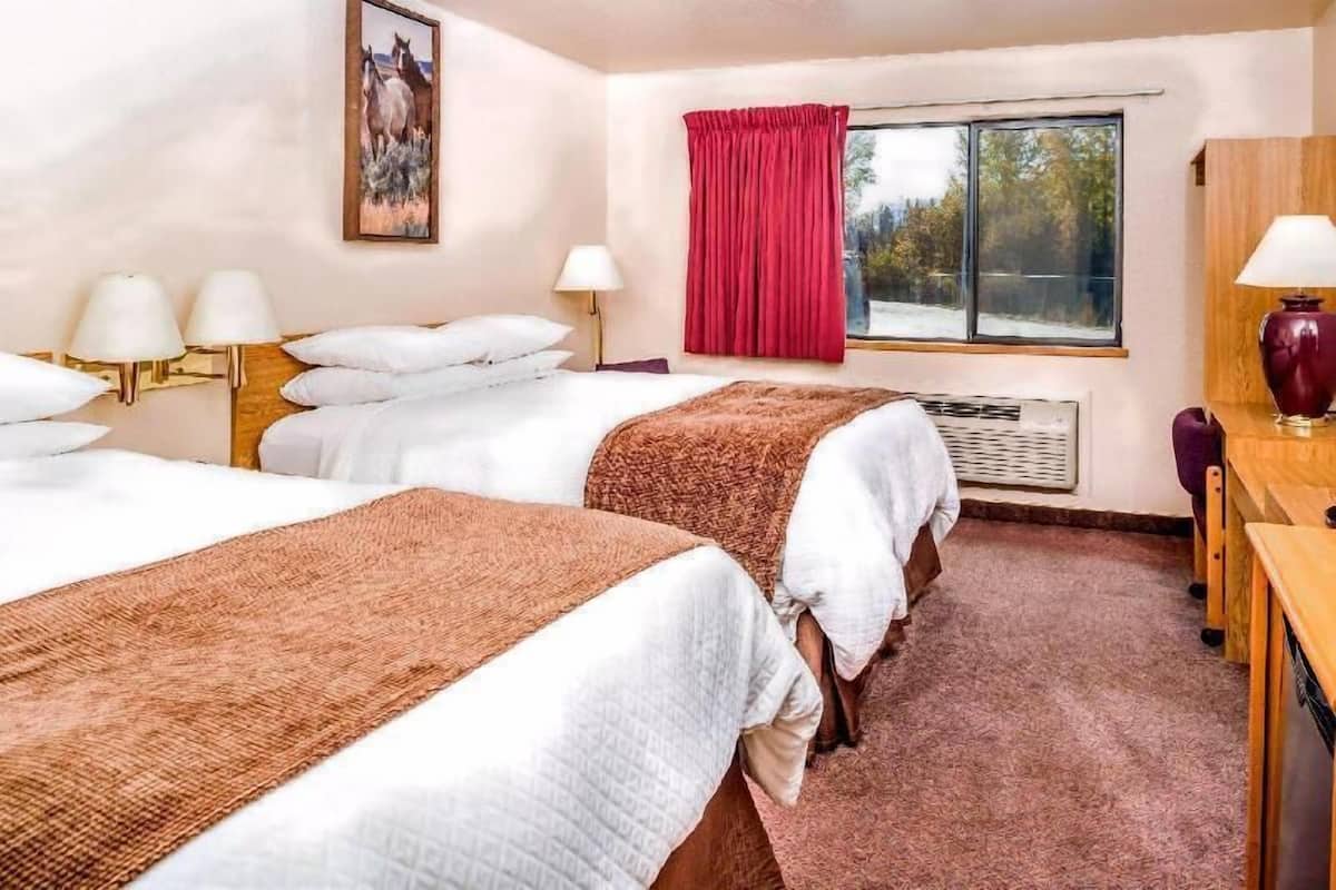 A hotel room with two beds, a desk, a chair, a window with a red curtain, and a painting on the wall.