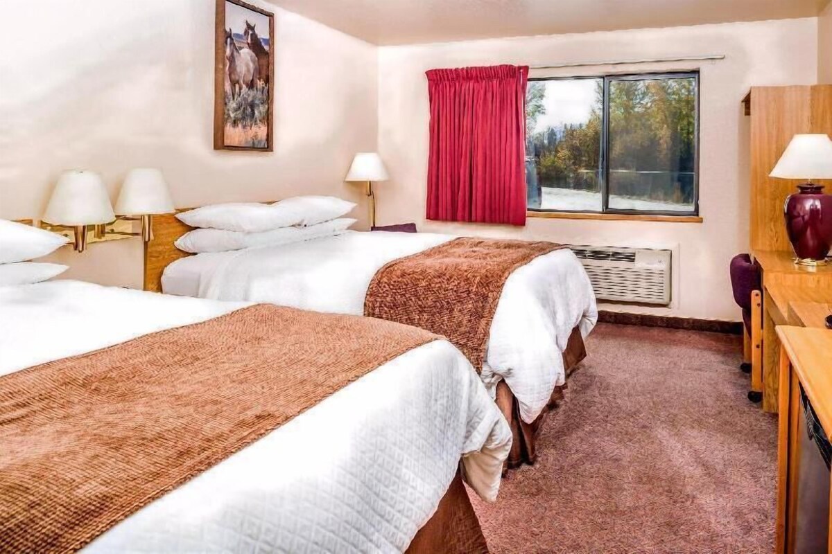 A hotel room with two beds, a desk, a chair, a window with a red curtain, and a painting on the wall.