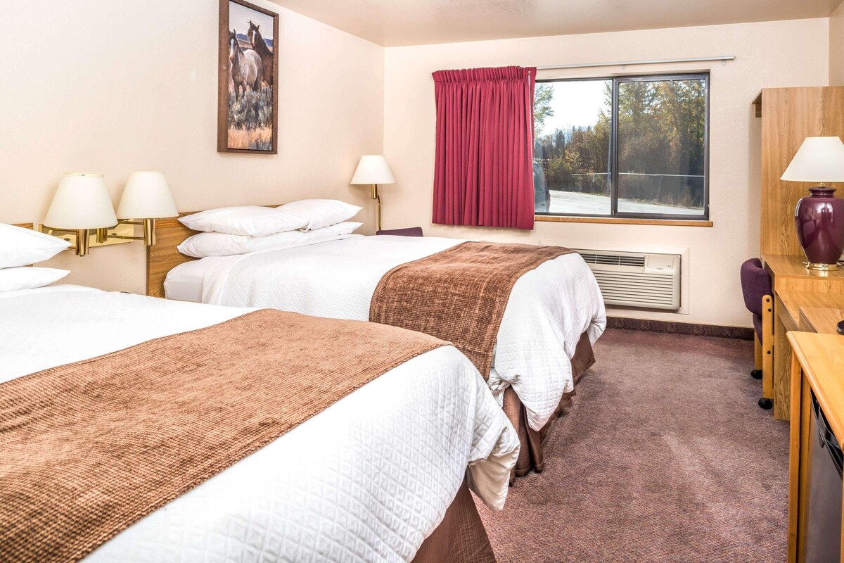 A hotel room with two beds, a desk, a chair, a window with red curtains, a painting on the wall, and a lamp.