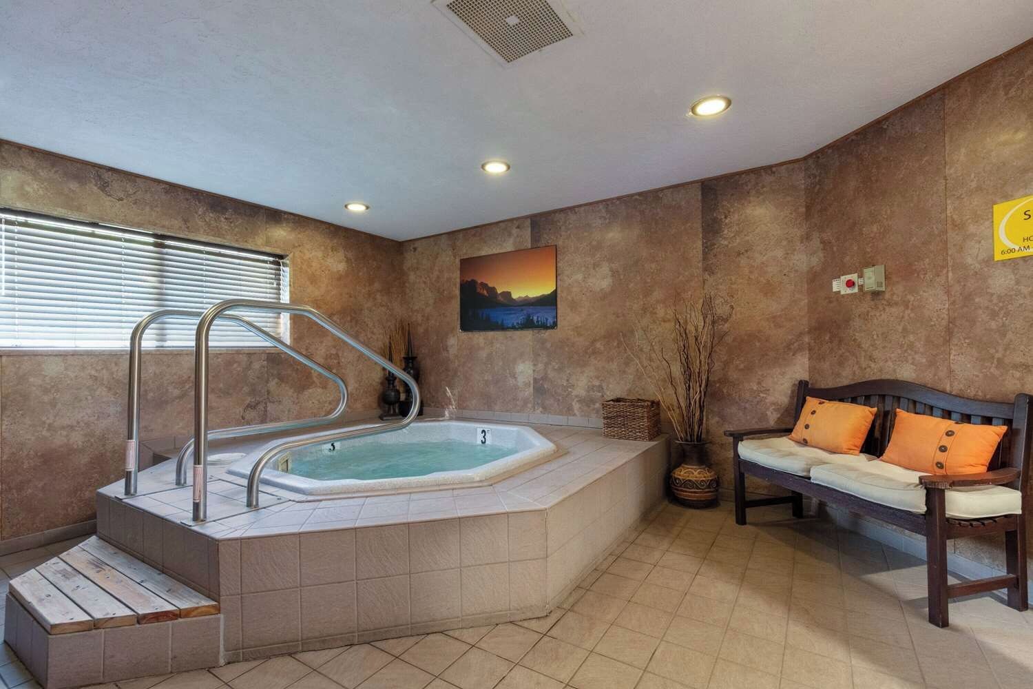 Un cuarto de spa con un jacuzzi, escalones de madera, un banco y una pintura de atardecer en la pared.