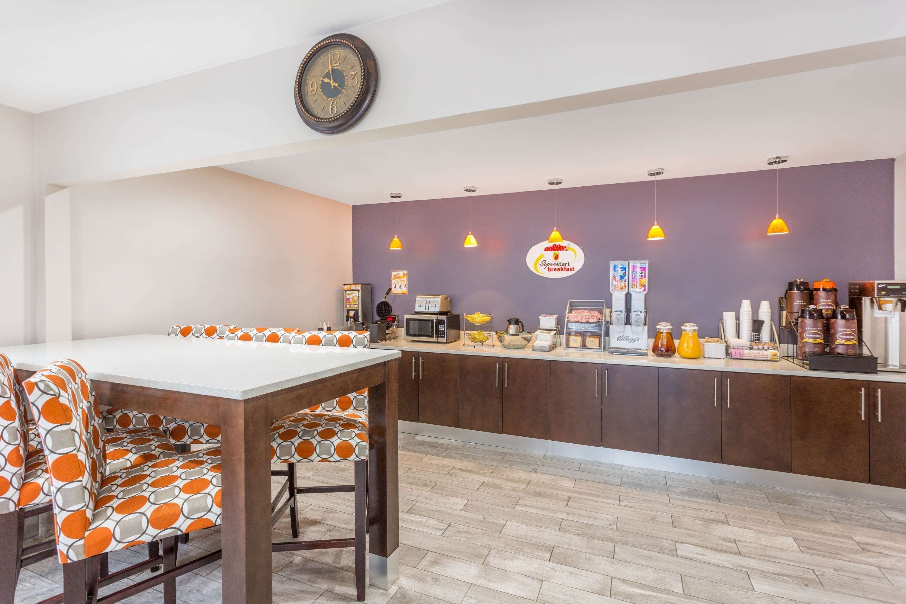 A modern kitchen with a dining area, featuring a white table, orange and gray patterned chairs, a clock, and various kitchen appliances and supplies.