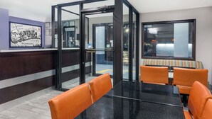 A modern reception area with a glass partition, a reception desk, and orange chairs around a black table. - Super 8 by Wyndham Locust Grove (Locust Grove)