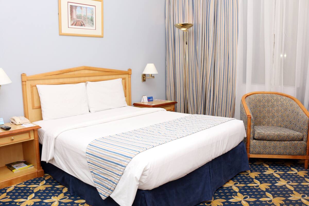 Une chambre d’hôtel avec un lit en bois, une chaise, une table de chevet avec un téléphone et une lampe.