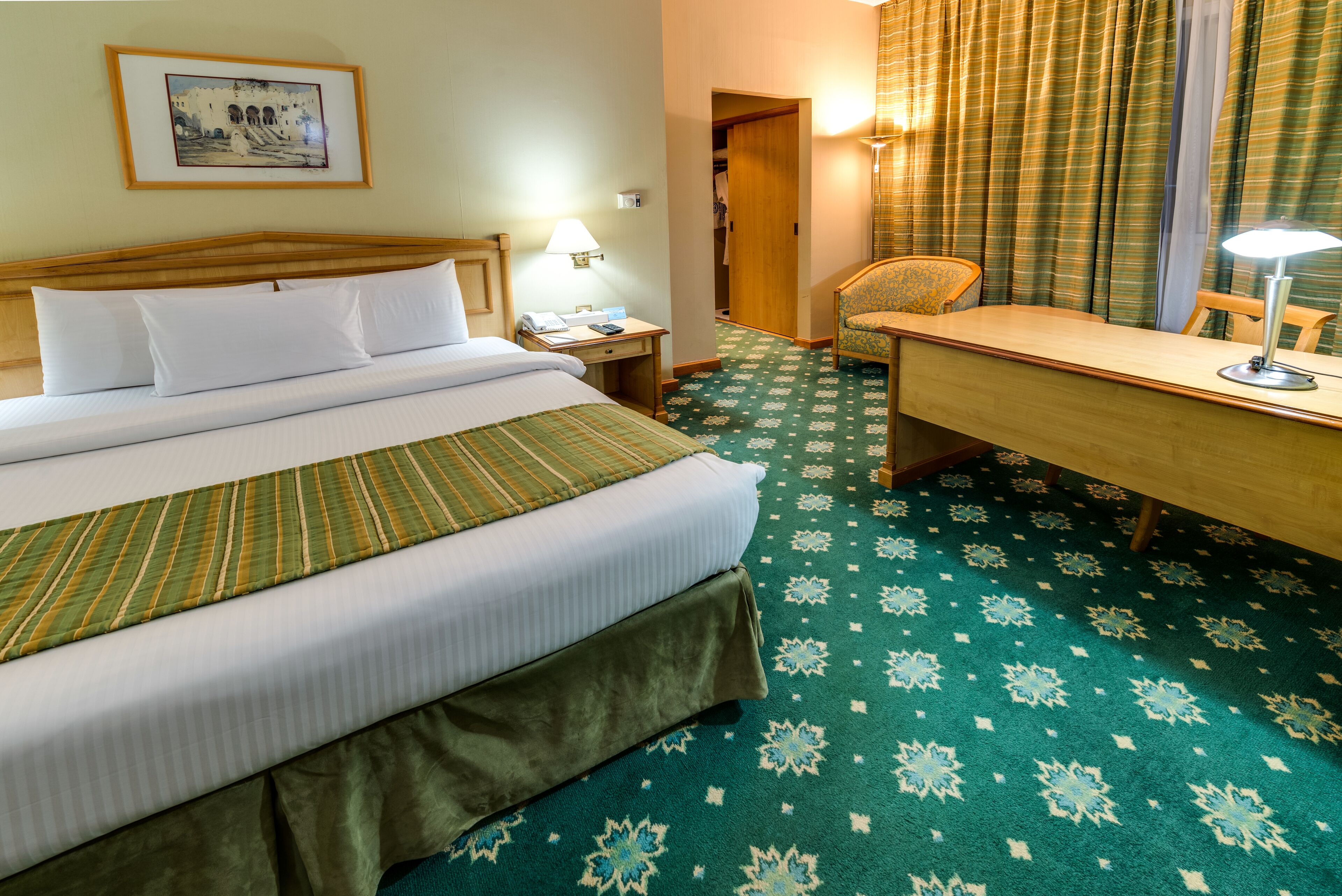A hotel room with a bed, a desk, a chair, a lamp, and a framed picture on the wall.