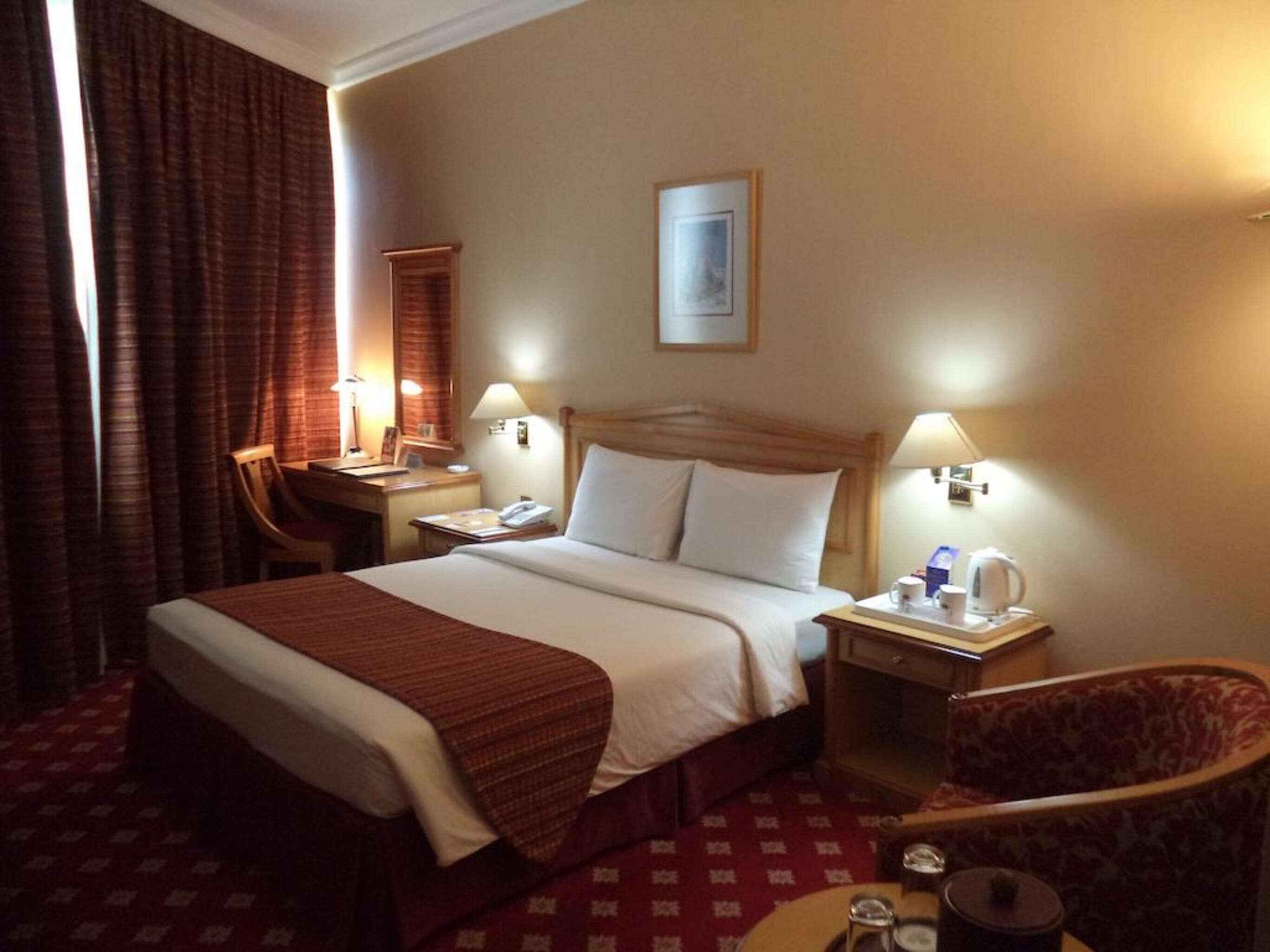 A hotel room with a bed, bedside tables, a desk, a chair, and a small table with a kettle and cups.