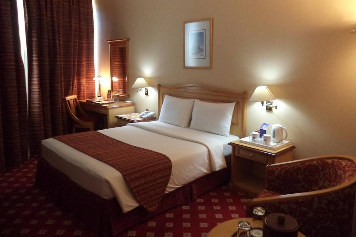 A hotel room with a bed, bedside tables, a desk, a chair, and a small table with a kettle and cups.