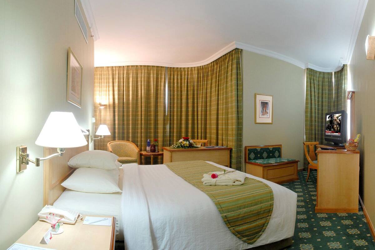Une chambre d’hôtel comprenant un lit, une table de chevet, une lampe, une chaise, un bureau, une télévision et des rideaux.