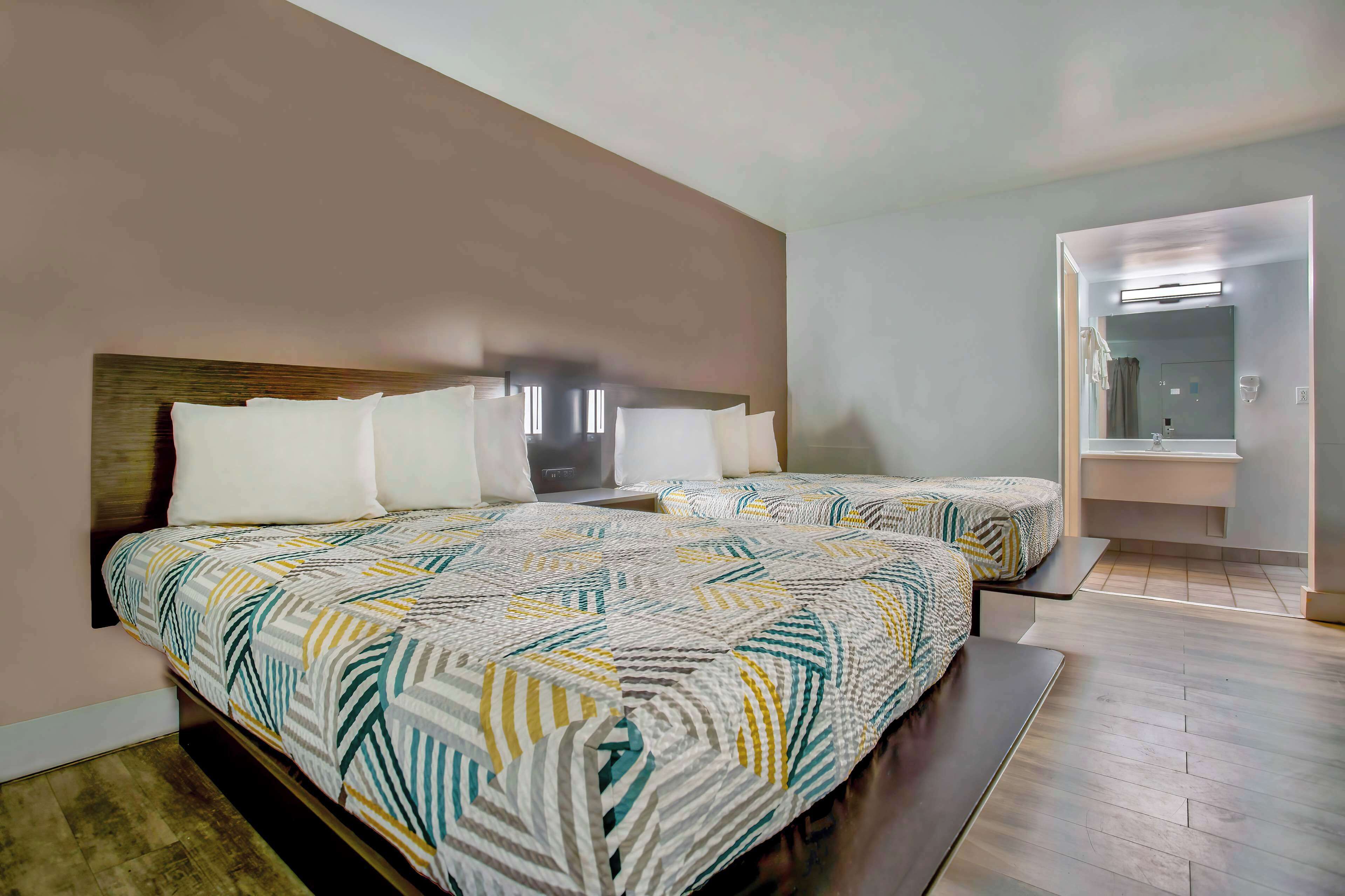 Habitación de hotel con dos camas, cabecera de madera, un espejo grande y un baño visible a través de una puerta abierta.