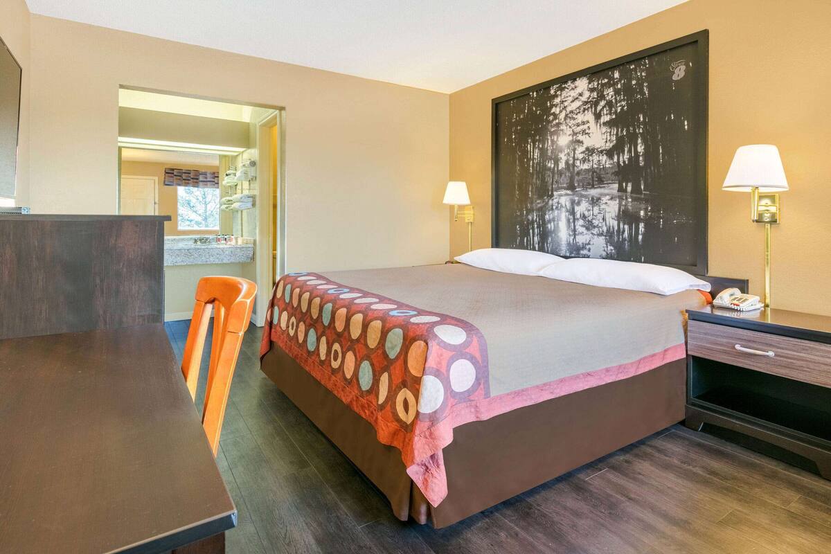 A hotel room with a bed, a desk, a chair, a nightstand, and a wall-mounted lamp.