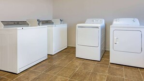 A laundry room with three white washing machines and a dryer, arranged in a row against a plain wall with brown tiled flooring.