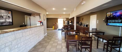 A hotel lobby with a reception desk, a clock, a TV screen, and a dining area with tables and chairs.