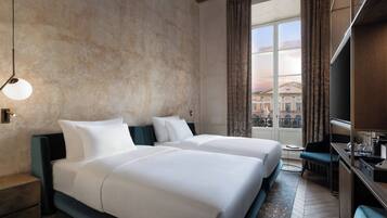 A hotel room with two beds, a desk, a chair, a TV, and a large window with curtains.