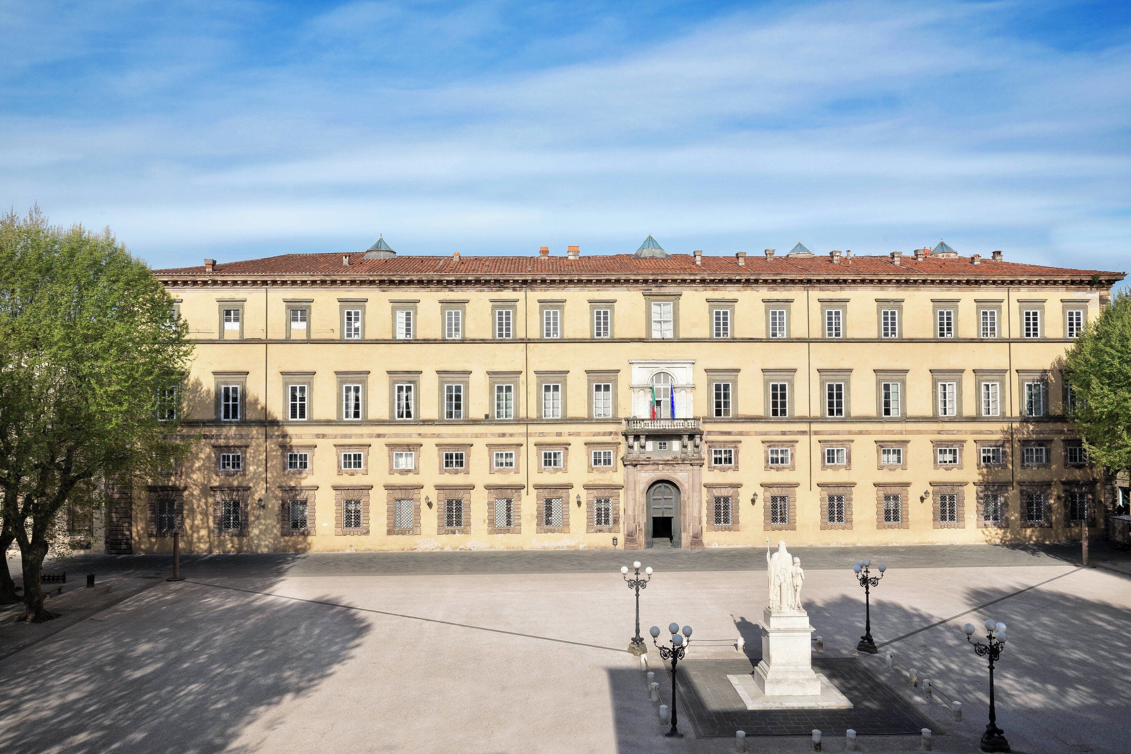 A large, historic building with symmetrical architecture, a central entrance, and a statue in the courtyard.