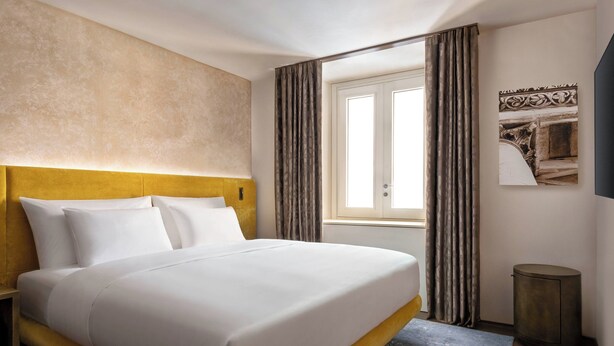 A hotel room with a large bed, a headboard, a window with curtains, a small trash bin, and a wall-mounted artwork. - Grand Universe Lucca, Autograph Collection (Lucca)