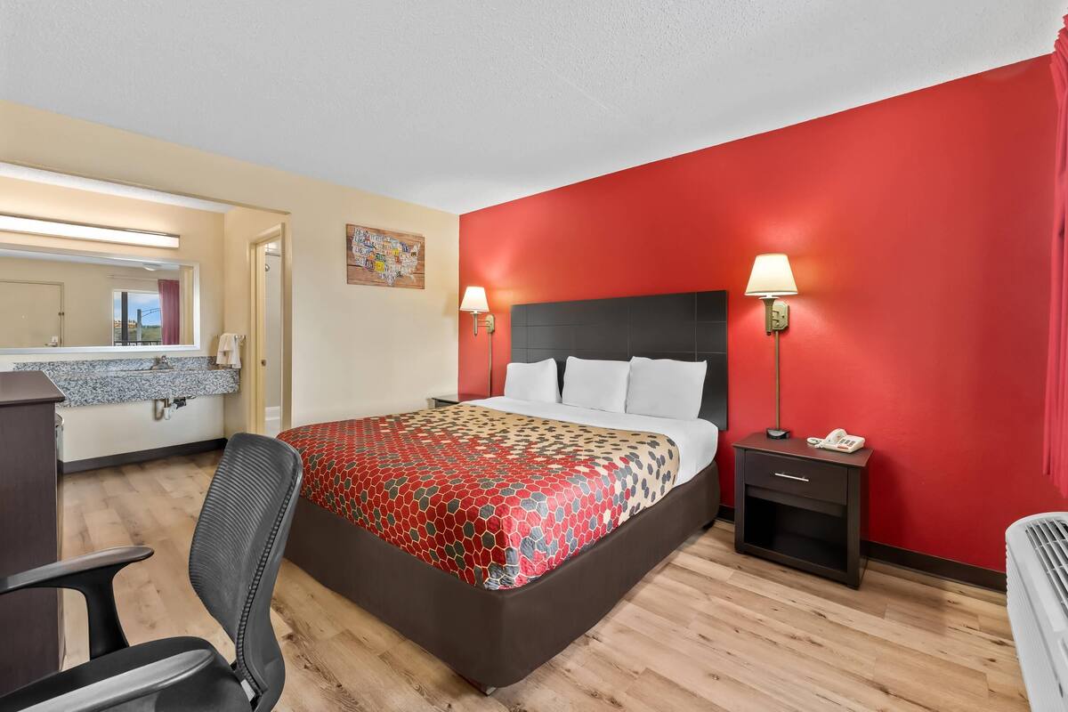 Una habitación de hotel con cama, mesita de noche, lámpara, silla y un baño visible a través de una puerta abierta.